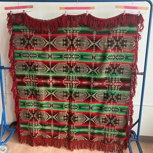 May include: A colorful blanket with a geometric pattern in red, green, brown, and tan. The blanket has a fringed edge in a deep red color. The blanket is displayed on a blue metal rack.
