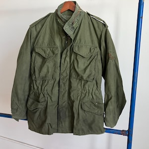 May include: Olive green military-style jacket with multiple front pockets, a stand-up collar, and a drawstring waist. The jacket is hanging on a wooden hanger. The jacket has a button-up front and a patch on the upper arm.