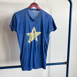 May include: A vintage blue t-shirt with a large, gold star design on the front. The short-sleeved shirt is hanging on a wooden hanger. The shirt has a crew neck and a slightly faded appearance.
