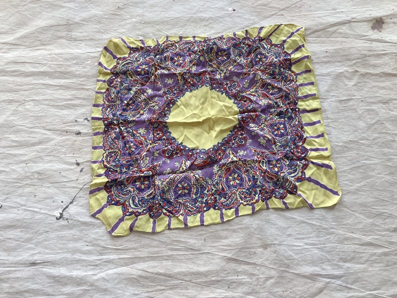 May include: A square silk scarf featuring a central yellow circle and a detailed purple and red paisley design. The border is yellow with vertical purple stripes. The scarf has a vintage look and a soft, worn texture.