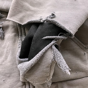 May include: A gray hooded sweatshirt with a distressed look, featuring several holes and frayed edges. The hood is pulled up and the sweatshirt is folded over itself, revealing a black fabric lining.