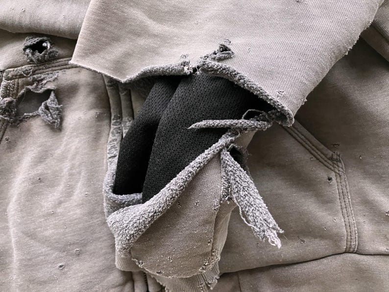 May include: A gray hooded sweatshirt with a distressed look, featuring several holes and frayed edges. The hood is pulled up and the sweatshirt is folded over itself, revealing a black fabric lining.