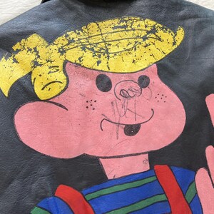 Op de afbeelding: Zwarte leren jas met een cartoonfiguur. Het personage heeft geel haar en een roze gezicht, draagt een blauw-groen gestreepte top met rode bretels. Het personage steekt zijn middelvinger op.