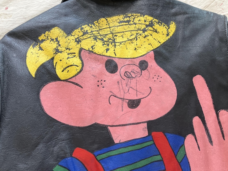 Op de afbeelding: Zwarte leren jas met een cartoonfiguur. Het personage heeft geel haar en een roze gezicht, draagt een blauw-groen gestreepte top met rode bretels. Het personage steekt zijn middelvinger op.