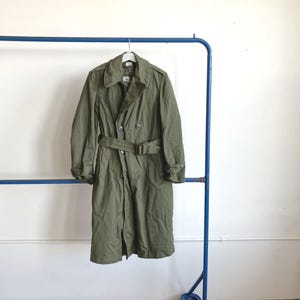 May include: Olive green trench coat with a double-breasted design, a belt, and a collar. The coat is hanging on a blue clothing rack against a white wall. The coat is a classic style.