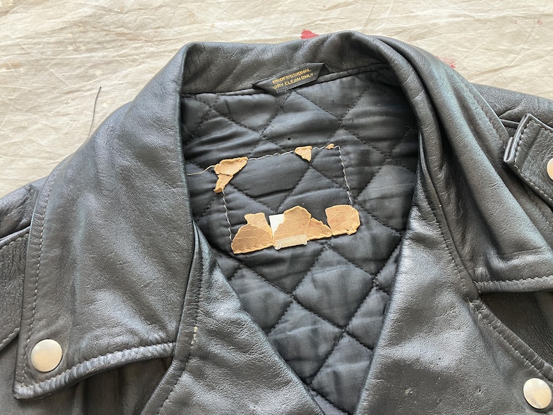 Op de afbeelding: Close-up van een zwarte leren jas met een gewatteerde voering. De jas heeft een reverskraag, zilveren drukkers en een label met de tekst "Professional Dry Cleaning". Stukjes karton zijn zichtbaar op de voering.