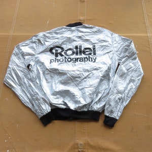 Puede incluir: Una chaqueta plateada, arrugada, con cremallera y ribetes negros, con las palabras "Rollei photography" impresas en la espalda.