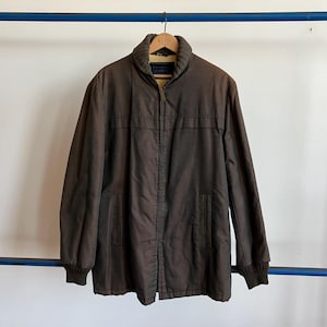 Puede incluir: Chaqueta marrón con cremallera y cuello. La chaqueta tiene dos bolsillos delanteros y puños de canalé. La chaqueta está colgada de una percha de madera sobre un fondo blanco.