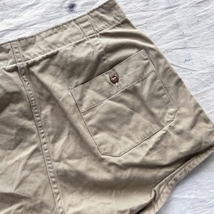 May include: Close-up of tan shorts with a buttoned pocket. The shorts have a high waist and visible stitching details. The fabric appears to be a sturdy cotton or linen blend, suitable for casual wear.