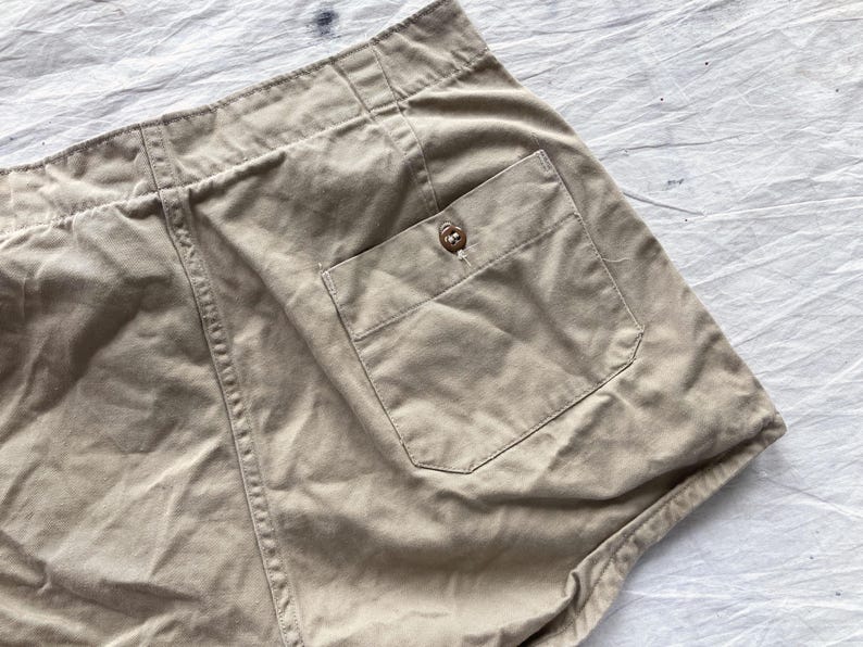 May include: Close-up of tan shorts with a buttoned pocket. The shorts have a high waist and visible stitching details. The fabric appears to be a sturdy cotton or linen blend, suitable for casual wear.