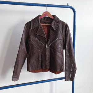 May include: A vintage brown leather jacket with a collared neckline and a zipper closure. The jacket has a fitted silhouette and features two zippered pockets on the front. It is hanging on a pink wooden hanger.
