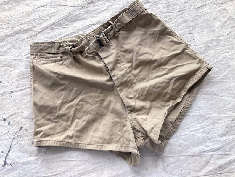 May include: Khaki-colored shorts with a belt and buckle. The shorts have a classic design with a front pocket and a belt loop. The fabric appears to be a durable cotton blend. The shorts are laid flat on a white surface.
