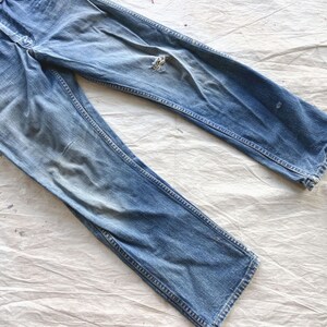Peut inclure: Jean bleu d&eacute;lav&eacute; avec un aspect us&eacute;. Le jean a une conception classique &agrave; cinq poches, une fermeture &agrave; bouton et &agrave; glissi&egrave;re, et une coupe droite. Il y a une d&eacute;chirure sur le genou droit. Le jean est pos&eacute; &agrave; plat sur une surface blanche.