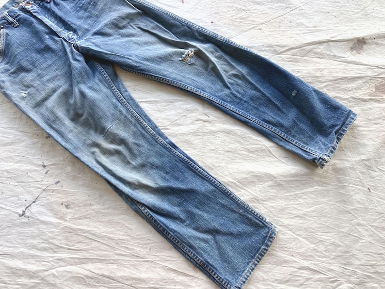Peut inclure: Jean bleu d&eacute;lav&eacute; avec un aspect us&eacute;. Le jean a une conception classique &agrave; cinq poches, une fermeture &agrave; bouton et &agrave; glissi&egrave;re, et une coupe droite. Il y a une d&eacute;chirure sur le genou droit. Le jean est pos&eacute; &agrave; plat sur une surface blanche.