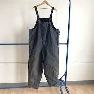 May include: Dark gray work overalls with adjustable straps and a bib. The overalls are hanging on a wooden hanger, suspended from a blue metal rack. The garment appears worn, with some discoloration.