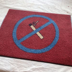 May include: A rectangular red doormat with a blue circle and a crossed-out cigarette symbol. The cigarette is beige and brown. The mat has a black border and a textured surface.
