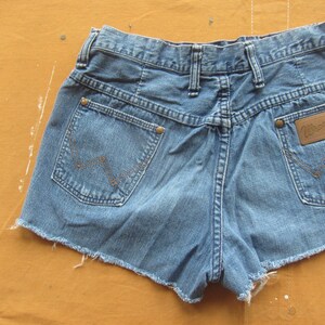 Op de afbeelding: Een lichtblauwe denim short met rafelige randen. De short heeft een bruin leren patch met het woord "Wrangler" op de rechter achterzak.