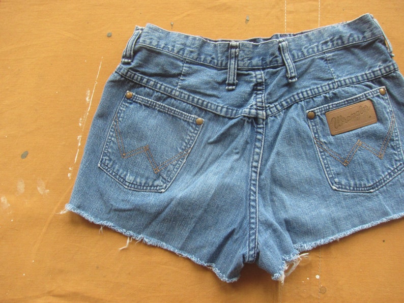 Op de afbeelding: Een lichtblauwe denim short met rafelige randen. De short heeft een bruin leren patch met het woord "Wrangler" op de rechter achterzak.