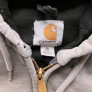 May include: Gray Carhartt hoodie with a black hood and a zipper closure. The hoodie has a label that says "Carhartt" and "XL Tall Made in Mexico".