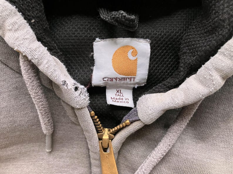 May include: Gray Carhartt hoodie with a black hood and a zipper closure. The hoodie has a label that says "Carhartt" and "XL Tall Made in Mexico".