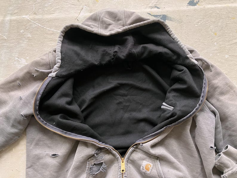 May include: A gray hooded sweatshirt with a black fleece lining. The sweatshirt has a zipper closure and a small logo patch on the front.