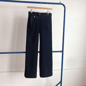 May include: Navy blue wide-leg pants hanging on a wooden hanger. The pants have a high waist with decorative buttons and side pockets. The fabric appears to be a medium-weight material, suitable for various seasons.