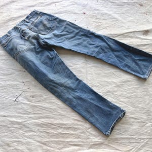 Peut inclure: Jean bleu d&eacute;lav&eacute; avec une conception classique &agrave; cinq poches. Le jean est pos&eacute; &agrave; plat sur une surface blanche textur&eacute;e, mettant en valeur les d&eacute;tails us&eacute;s et les subtiles variations de couleur. Le jean semble &ecirc;tre de longueur standard.