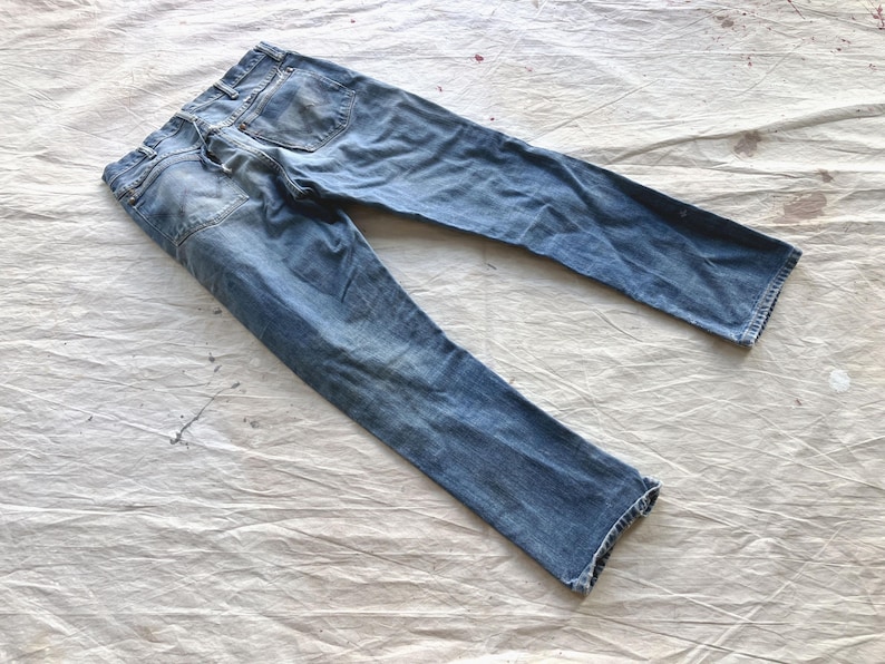Peut inclure: Jean bleu d&eacute;lav&eacute; avec une conception classique &agrave; cinq poches. Le jean est pos&eacute; &agrave; plat sur une surface blanche textur&eacute;e, mettant en valeur les d&eacute;tails us&eacute;s et les subtiles variations de couleur. Le jean semble &ecirc;tre de longueur standard.