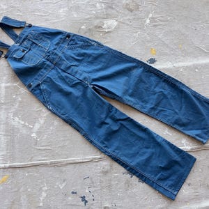 Könnte beinhalten: Eine blaue Denim-Latzhose mit verstellbaren Trägern. Die Latzhose hat einen klassischen Arbeitsstil und ist aus strapazierfähigem Stoff gefertigt.