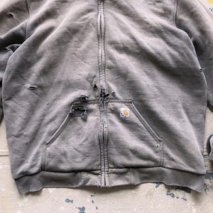 May include: A gray zip-up hoodie with a distressed look. The hoodie has a large pocket on the front and a small logo patch on the pocket. The hoodie has a zipper closure and a hood.