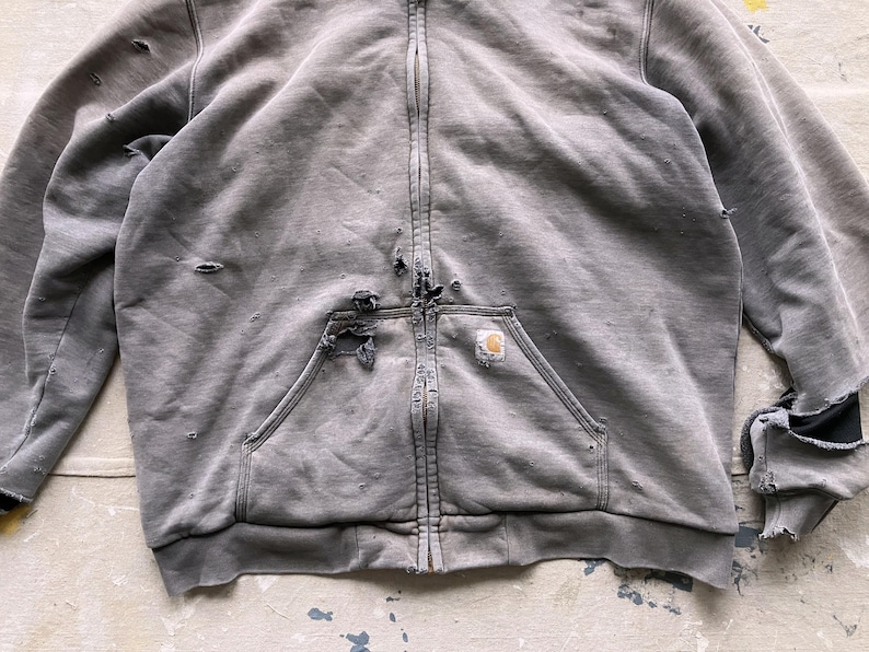 May include: A gray zip-up hoodie with a distressed look. The hoodie has a large pocket on the front and a small logo patch on the pocket. The hoodie has a zipper closure and a hood.