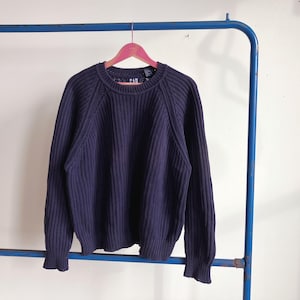 May include: A navy blue ribbed knit jumper with a crew neck, hanging on a pink hanger. The jumper has long sleeves and a ribbed hem and cuffs. The brand "GAP" is visible on the label.