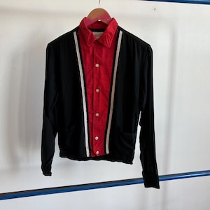 May include: Black long-sleeve shirt with a red front panel and white stripes. The shirt has a collared neckline and button-up closure. The shirt is hanging on a wooden hanger.