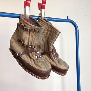 Puede incluir: Un par de botas de tela acolchada color canela con ribetes y suelas de cuero marrón oscuro. Las botas presentan una franja vertical verde oliva en la parte delantera, correas de cuero marrón con hebillas metálicas y remaches plateados.