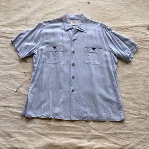 May include: Light blue short-sleeved shirt with a camp collar and two chest pockets. The shirt has a button-down front and a subtle vertical texture. The pockets have a dark blue triangle design. The shirt is laid flat on a neutral-toned surface.