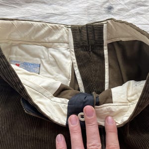 K&ouml;nnte beinhalten: Nahaufnahme des Hosenbunds einer braunen Cordhose. Die Hose hat einen Knopfverschluss, einen Rei&szlig;verschluss und ein wei&szlig;es Futter. Das Etikett lautet "OBERMEYER".