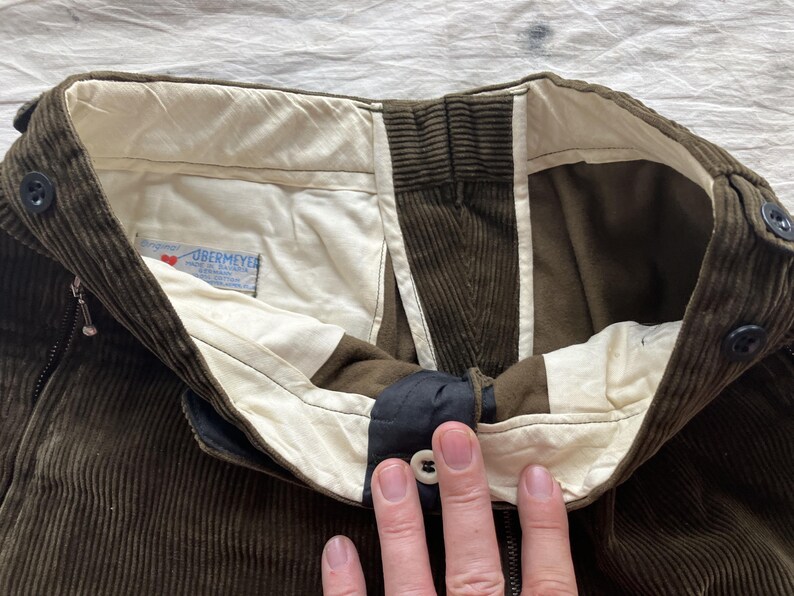 K&ouml;nnte beinhalten: Nahaufnahme des Hosenbunds einer braunen Cordhose. Die Hose hat einen Knopfverschluss, einen Rei&szlig;verschluss und ein wei&szlig;es Futter. Das Etikett lautet "OBERMEYER".