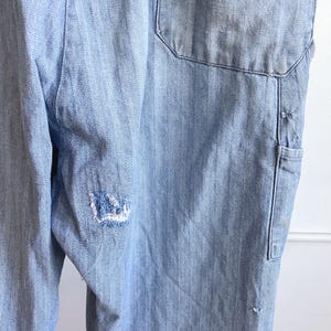 May include: Light blue denim overalls with a herringbone pattern. The overalls have a large pocket on the back and a small pocket on the side. There is a distressed area on the leg. The overalls are made of a durable denim material.