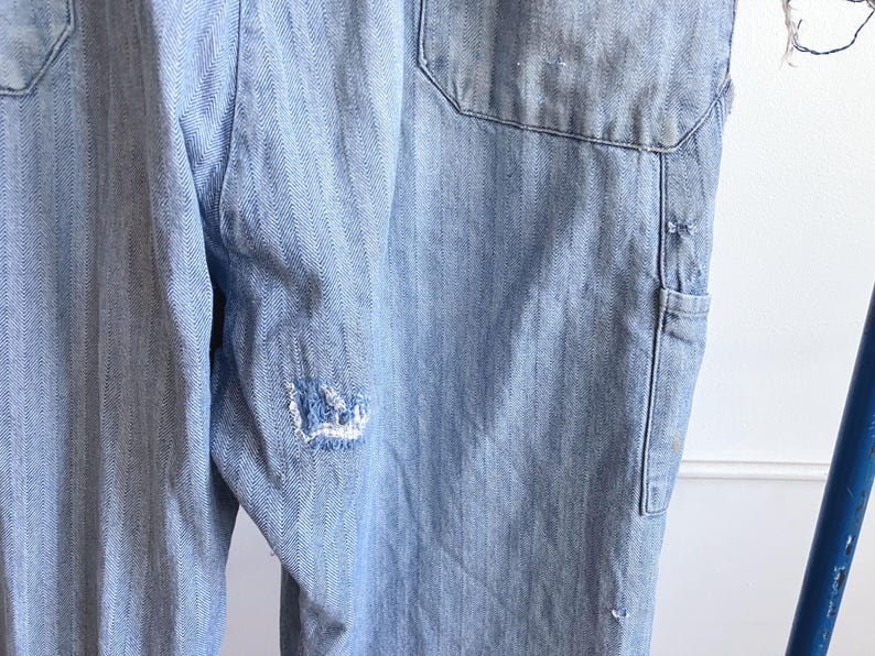May include: Light blue denim overalls with a herringbone pattern. The overalls have a large pocket on the back and a small pocket on the side. There is a distressed area on the leg. The overalls are made of a durable denim material.