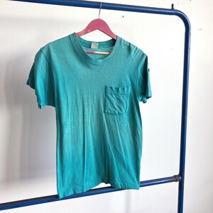 Könnte beinhalten: Ein türkisfarbenes T-Shirt mit Brusttasche hängt an einem rosa Kleiderbügel. Das Shirt hat einen Rundhalsausschnitt und kurze Ärmel. Das Kleidungsstück wird auf einem blauen Metallkleiderständer vor einer weißen Wand ausgestellt.