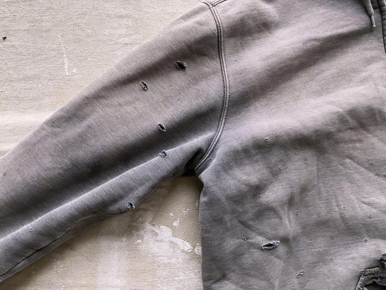 May include: A gray sweatshirt with several small holes throughout the fabric. The sweatshirt has a hood and long sleeves.