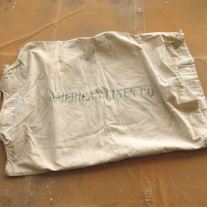 May include: A large, off-white canvas bag with a drawstring closure. The bag is made of heavy-duty canvas and has a faded, vintage look. The text "AMERICAN LINEN CO." is printed on the front of the bag.