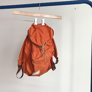 May include: A bright orange backpack featuring a rounded top flap, brown trim, and a beige drawstring closure. The bag includes a front pocket, brown adjustable shoulder straps, and a small white rectangular patch near the bottom edge.