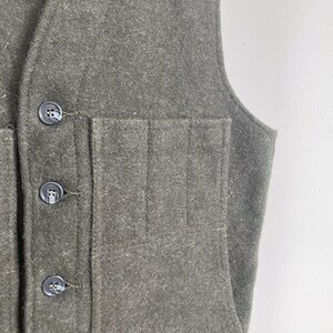 May include: A dark olive green vest made of a felted wool-like fabric. The garment features a front button closure with three dark circular buttons and a large patch pocket on the front panel that is divided into three vertical sections.
