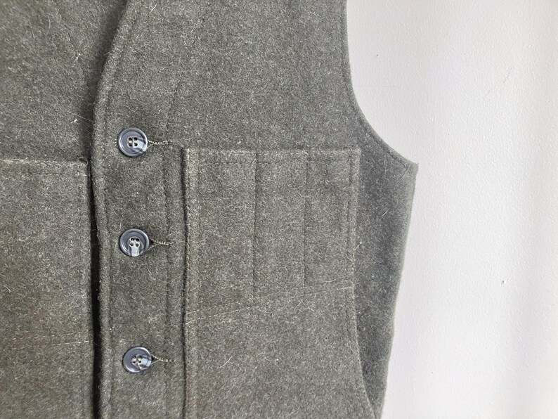 May include: A dark olive green vest made of a felted wool-like fabric. The garment features a front button closure with three dark circular buttons and a large patch pocket on the front panel that is divided into three vertical sections.