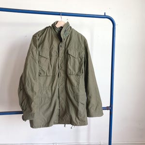 May include: An olive green military-style jacket with multiple pockets and a high collar. The jacket is hanging on a blue metal clothing rack. The jacket has a button-up front and a drawstring at the waist.