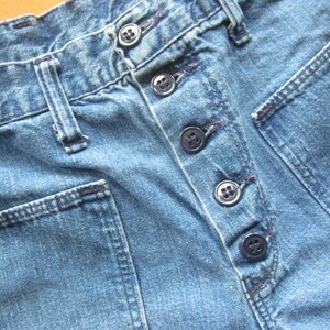 Op de afbeelding: Close-up van een paar blauwe denim jeans met een knoopsluiting en meerdere zakken. De jeans heeft een versleten en verbleekte uitstraling.