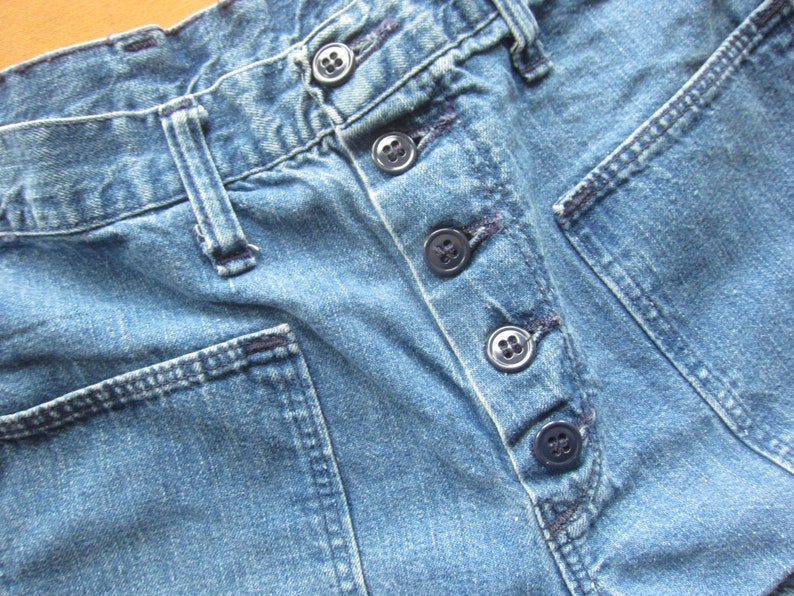 Op de afbeelding: Close-up van een paar blauwe denim jeans met een knoopsluiting en meerdere zakken. De jeans heeft een versleten en verbleekte uitstraling.