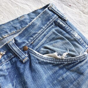 Peut inclure: Gros plan sur un jean en denim bleu d'aspect us&eacute;. Le jean pr&eacute;sente une conception classique &agrave; cinq poches, des rivets de couleur cuivre et une fermeture boutonn&eacute;e. La marque Wrangler est visible sur un bouton en cuivre.