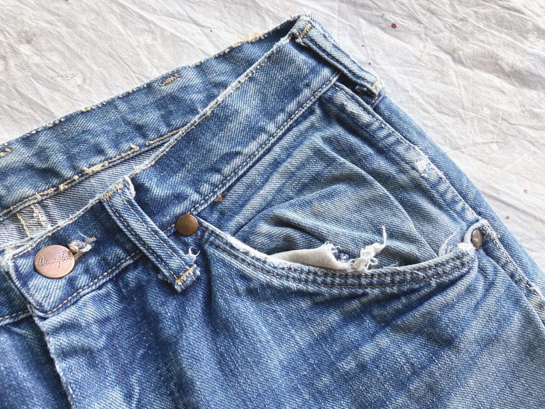 Peut inclure: Gros plan sur un jean en denim bleu d'aspect us&eacute;. Le jean pr&eacute;sente une conception classique &agrave; cinq poches, des rivets de couleur cuivre et une fermeture boutonn&eacute;e. La marque Wrangler est visible sur un bouton en cuivre.